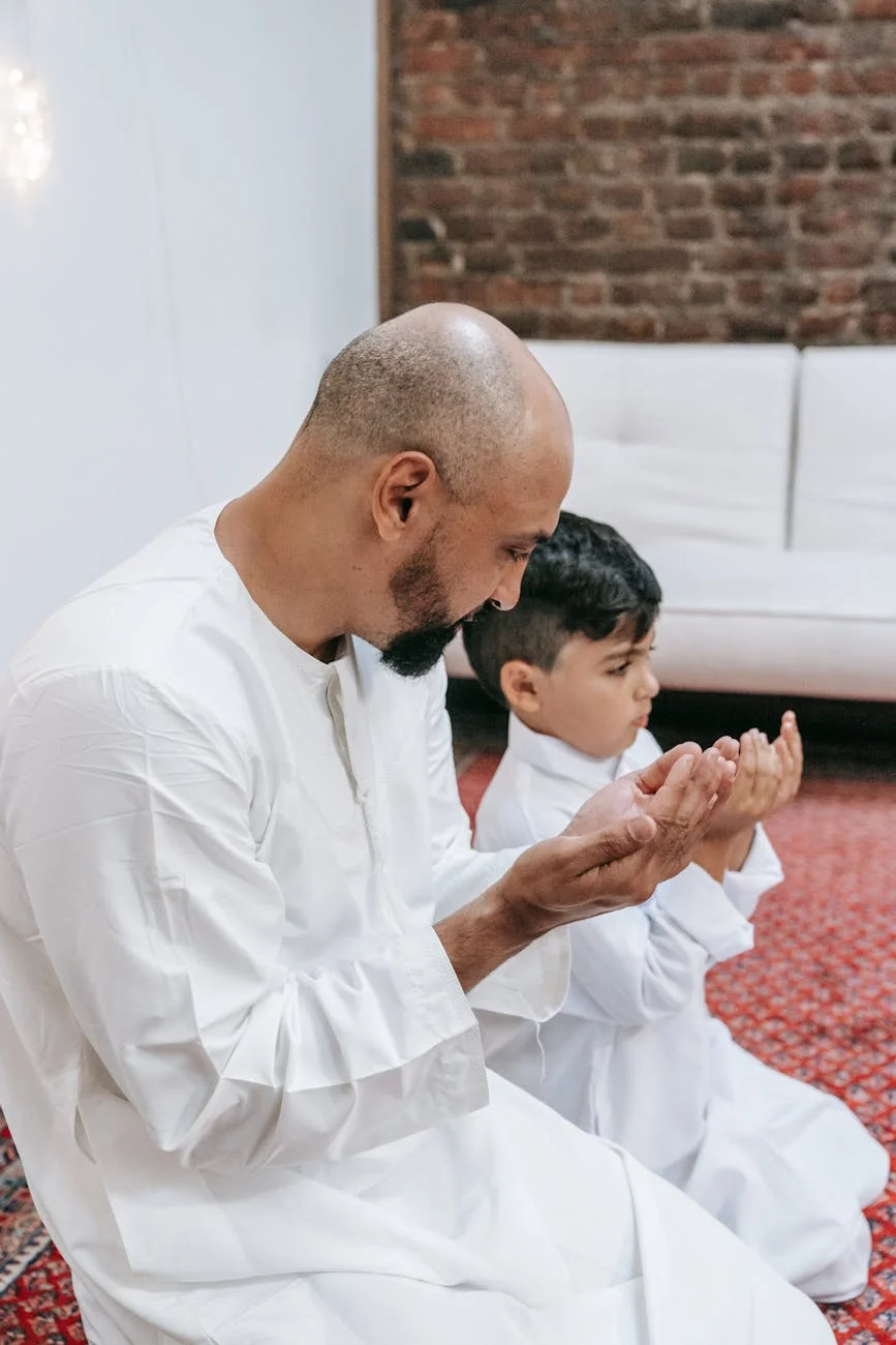 A bald, bearded man and young boy praying on a rug indoors, wearing traditional white clothing.