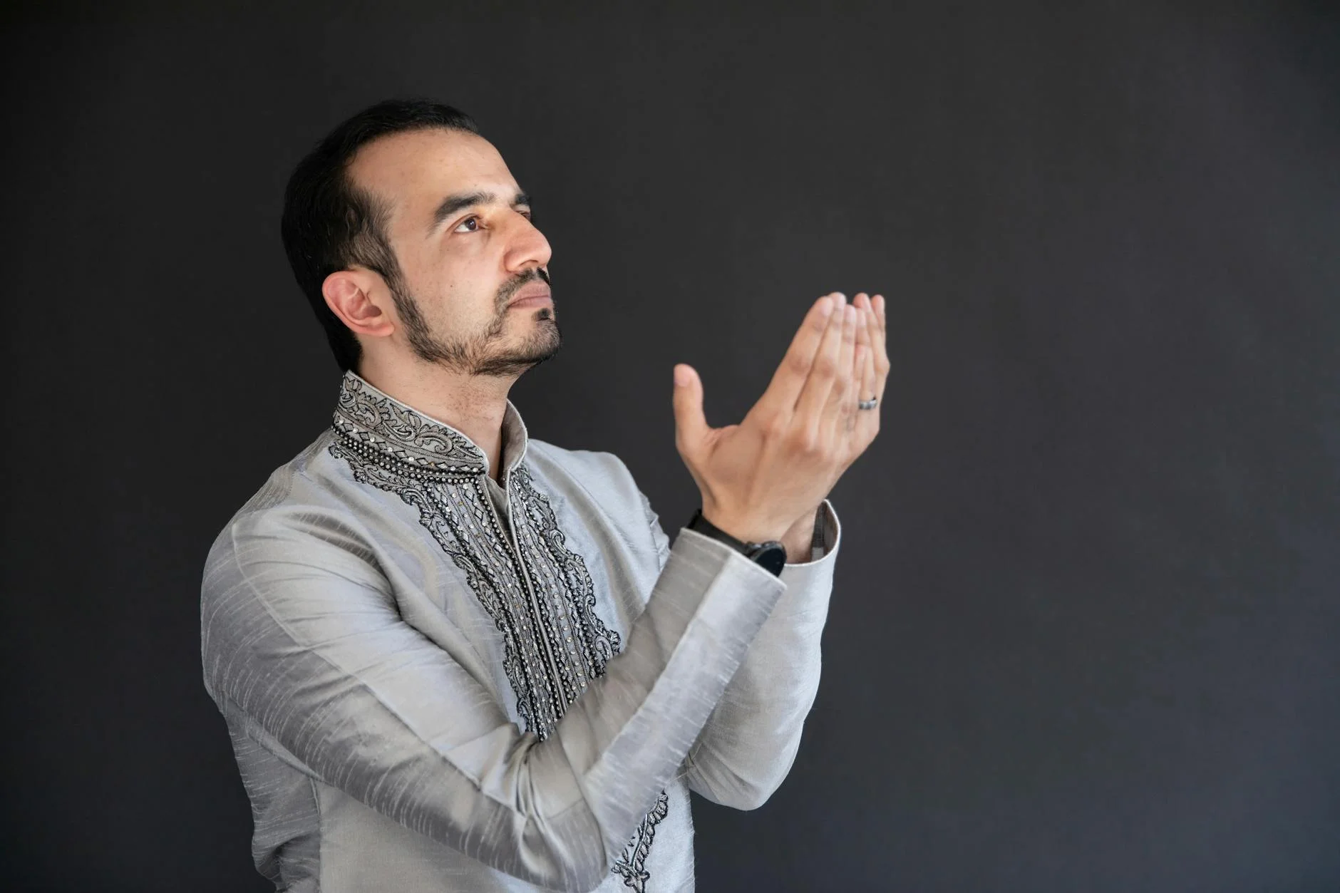 Middle Eastern man in traditional attire praying with hands raised against dark background.